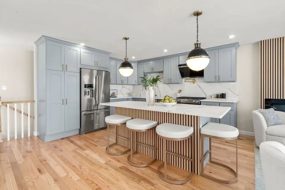 Quartz island with white stools, tall pantry cabinets and stainless refrigerator